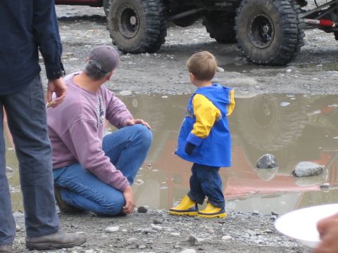 1990 Ford Bronco - A man and a child stand near a large tire in a muddy puddle, with another off-road vehicle in the background.