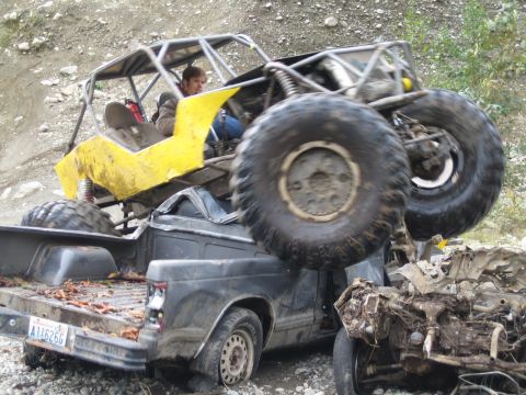 1990 Ford Bronco - An off-road buggy is precariously perched atop a pickup truck, with a driver in the buggy's cockpit.
