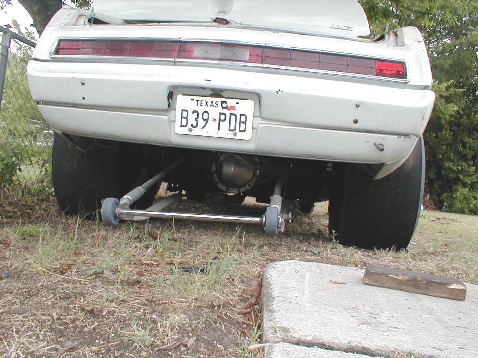 Rear of white coupe with Texas plate B39-PDB, wide rear tires and chrome bar under rear axle, parked on grass