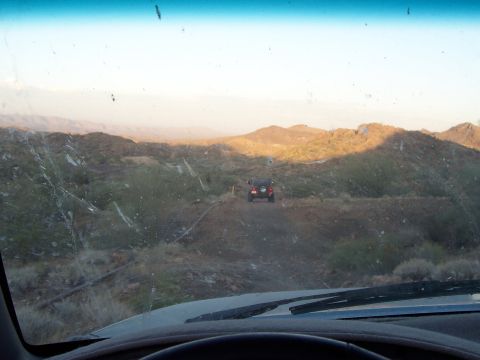 1996 Ford Bronco - Two off-road vehicles travel on a dirt road through a desert landscape at sunset.