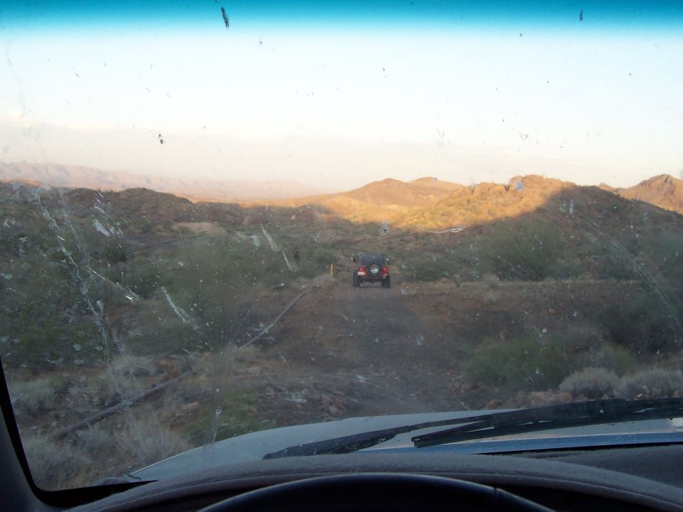 View from inside a vehicle looking at two off-road vehicles on a dirt path in a desert with hills