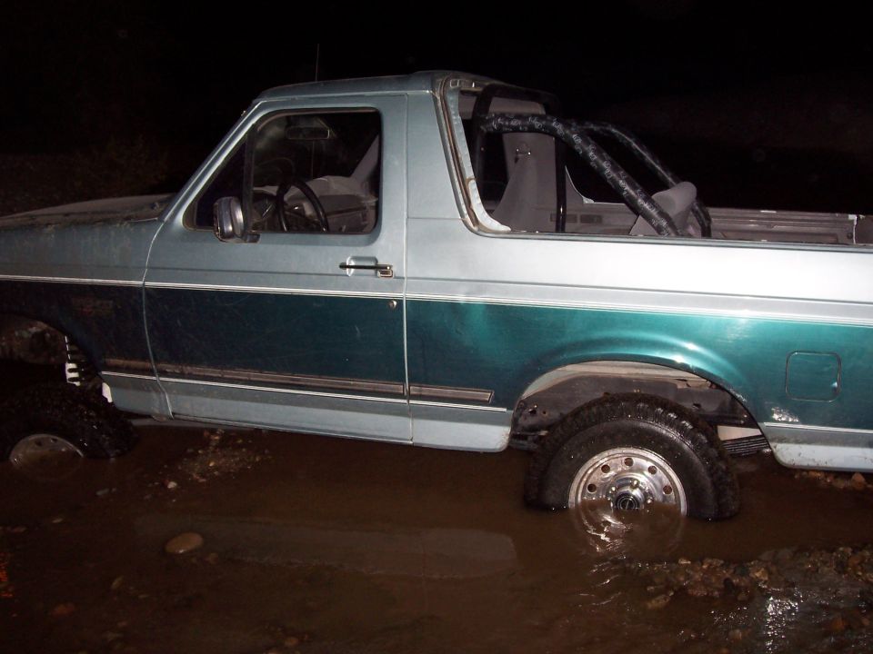 Side view of a 1996 Ford Bronco stuck in muddy water, with a roll cage visible inside
