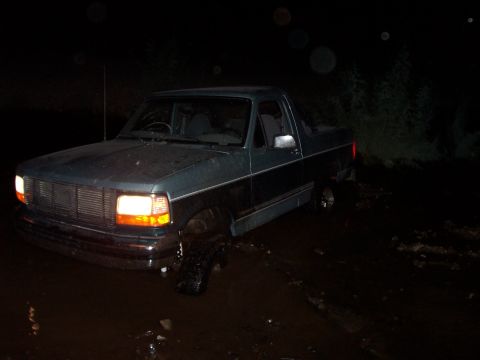 1996 Ford Bronco - A dark teal Ford F-150 is stuck in mud at night, with its headlights on.