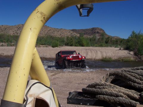1996 Ford Bronco - A red Jeep Wrangler navigates a riverbed under a clear blue sky with arid hills in the background.
