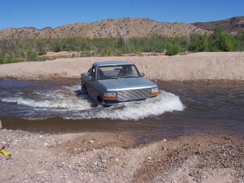 1996 Ford Bronco - A light blue pickup truck navigates through a shallow river, kicking up water as it goes.