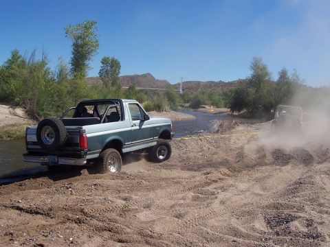 1996 Ford Bronco - Two off-road vehicles navigate a river crossing on a dusty trail under a clear blue sky.