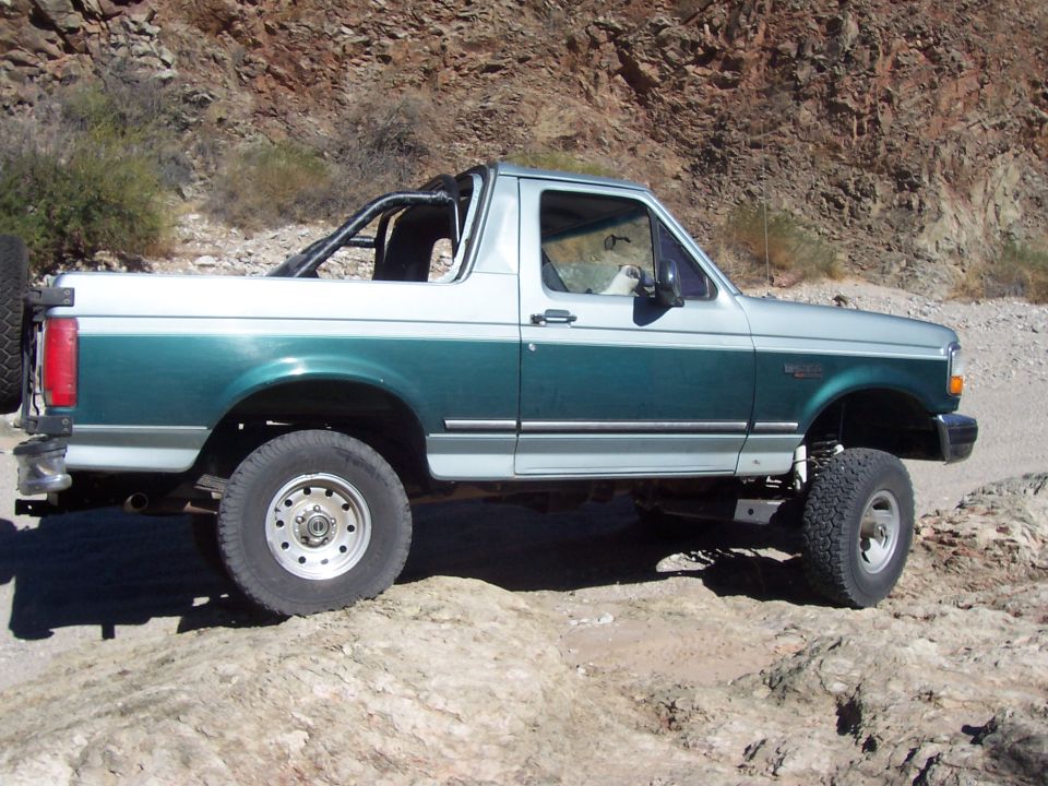 1996 Ford Bronco two-tone green and silver, lifted, off-road tires, spare tire on rear, driving on rocky terrain