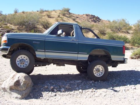 1996 Ford Bronco - A two-tone green and silver Ford Bronco is shown from the side, navigating a rocky desert landscape.