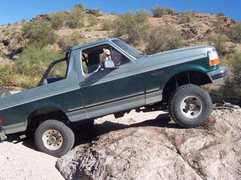 1996 Ford Bronco - A two-tone green Ford Bronco navigates a rocky off-road path with a dog visible in the driver's seat.