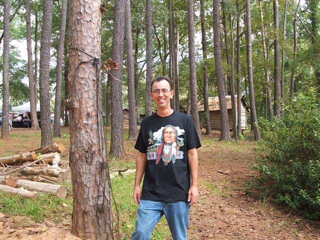 Man wearing a t-shirt with a Native American chief graphic stands in a wooded area with cabins in the background. "SUPERMOTORS.NET" logo in lower right