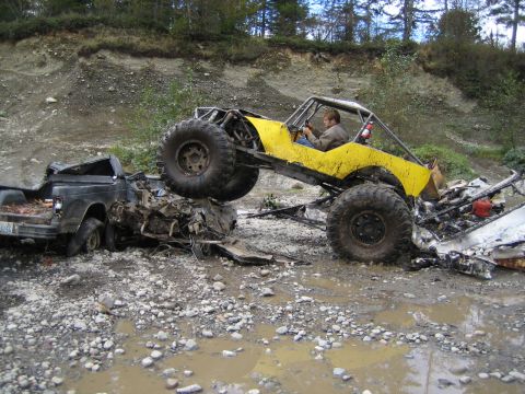 1990 Ford Bronco - A yellow off-road buggy is positioned precariously on top of a heavily damaged black pickup truck in a debris-filled, muddy area.