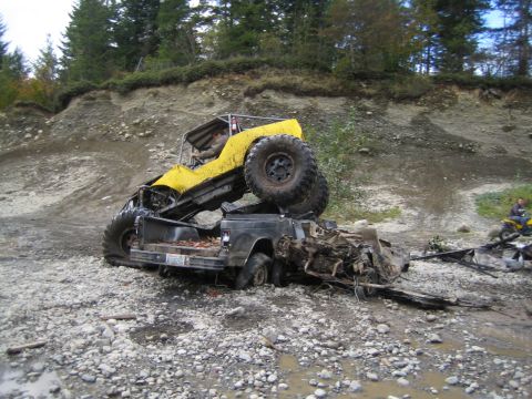 1990 Ford Bronco - A yellow off-road vehicle is precariously balanced on a heavily damaged black pickup truck in a rocky, outdoor environment.