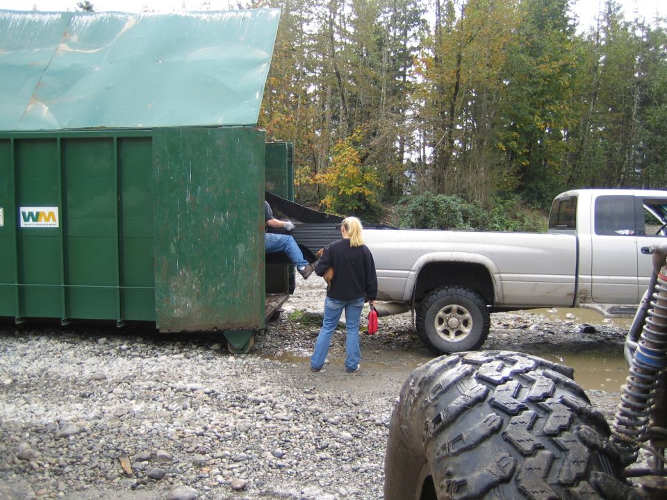 Silver pickup truck parked next to a green dumpster, with people loading it. Another vehicle's tire is in the foreground