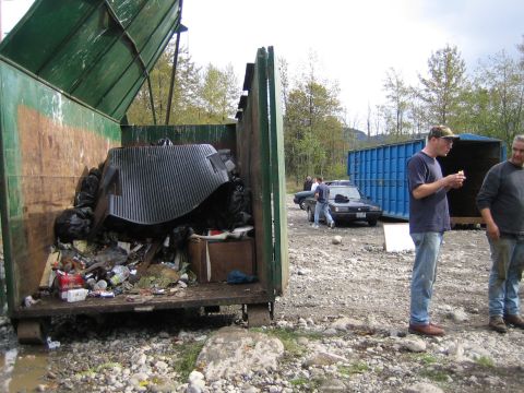 1990 Ford Bronco - A dumpster is overflowing with garbage, while two men stand near a car and a large blue container in the background.