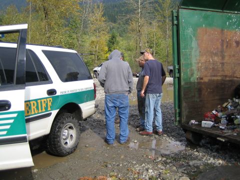 1990 Ford Bronco - A Sheriff SUV is parked next to a dumpster with several people standing nearby in a muddy outdoor scene.