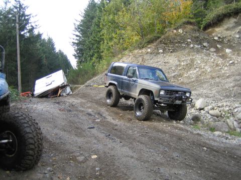 1990 Ford Bronco - Ford Bronco tows a wrecked trailer off-road, with another vehicle's tire in view.