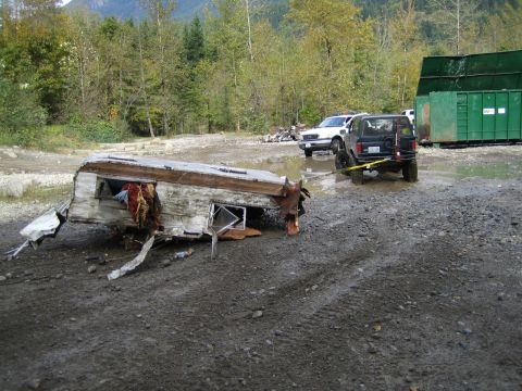 1990 Ford Bronco - A black SUV tows a heavily damaged trailer through a muddy area, with a white truck and green dumpsters in the background.