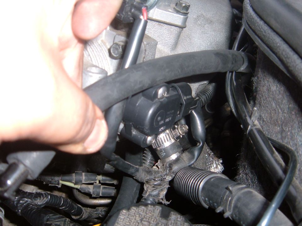 Close-up of a hand near a black sensor attached to hoses and wiring in an engine bay