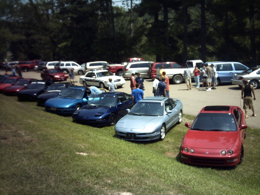 Several cars parked on grass, including a silver sports car and a red sports car in the foreground. Other vehicles and people are in the background
