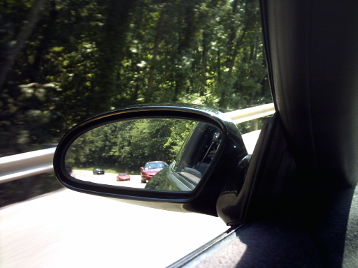 View of three cars on a road through a car's side mirror, with trees lining the road