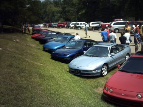 1995 Ford Probe - A gathering of cars parked on a grassy lot, with trees and people visible in the background.
