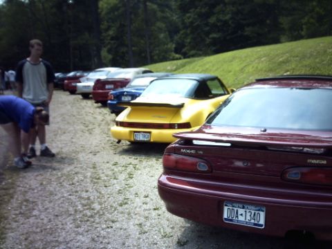 1995 Ford Probe - A line of cars parked on a gravel lot with a grassy hill and trees in the background.
