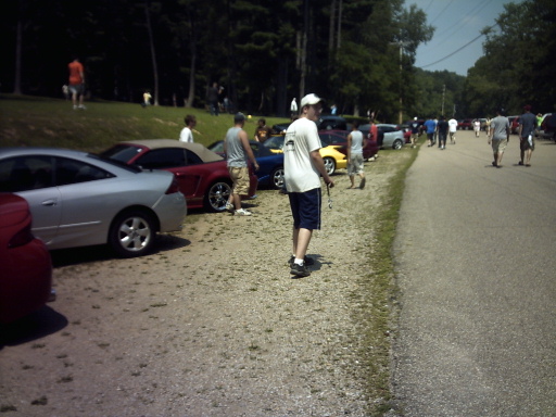 Several cars parked on a gravel and asphalt area next to a road with people standing around