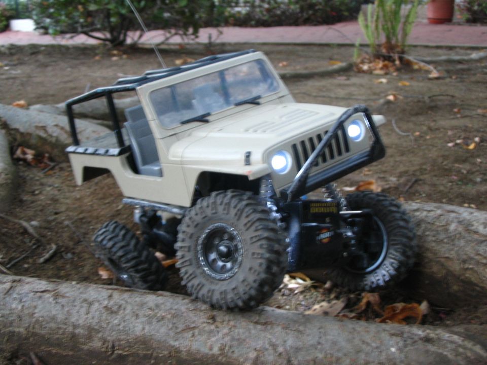 Tan RC Jeep crawler on a log over dirt, roll cage, LED lights and beadlock mud tires