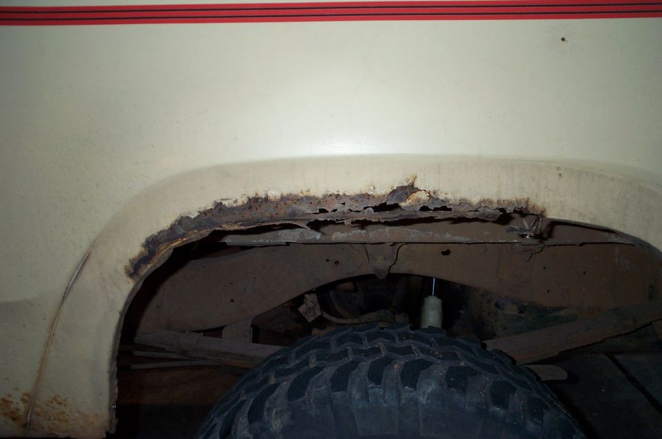 close-up of heavily rusted rear wheel well on a cream-colored vehicle with red trim above