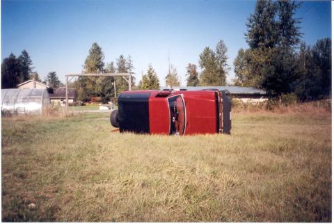 1989 Ford Bronco - A red and black SUV lies on its side in a field, with a greenhouse and buildings in the background.