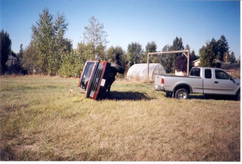 1989 Ford Bronco - A red SUV, identified as a 1989 Ford Bronco, lies on its side in a grassy field next to a silver pickup truck.