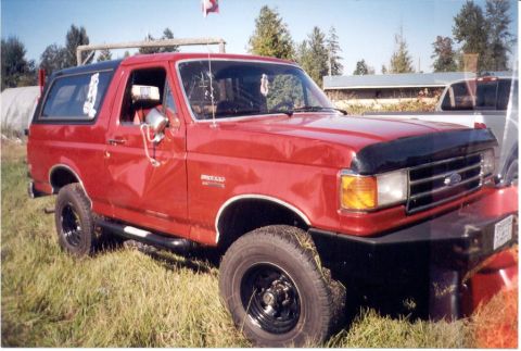 1989 Ford Bronco - This red Ford Bronco is parked in a grassy area with a second vehicle visible in the background.