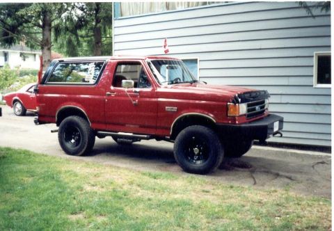 1989 Ford Bronco - A red Ford Bronco is parked in a driveway next to a house, with another red car visible in the background.