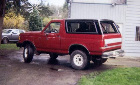 1989 Ford Bronco - Rear three-quarter view of a red full-size Ford Bronco parked on a wet driveway next to a white garage, with a white roof and white two-ton…
