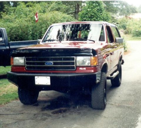 1989 Ford Bronco - Front three-quarter view of a red full-size Ford Bronco parked on a wet driveway, with a black hood bra, black front bumper, blue Ford oval…
