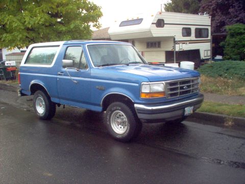 1992 Ford Bronco - A blue 1992 Ford Bronco with a white top is parked on a wet street next to a camper trailer.