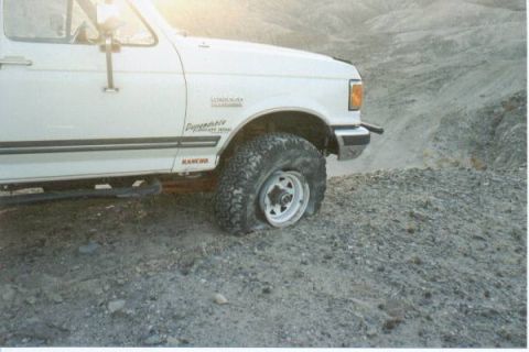 1990 Ford Bronco - A white 1990 Ford Bronco is pictured with a flat tire while off-roading on a rocky, barren landscape.