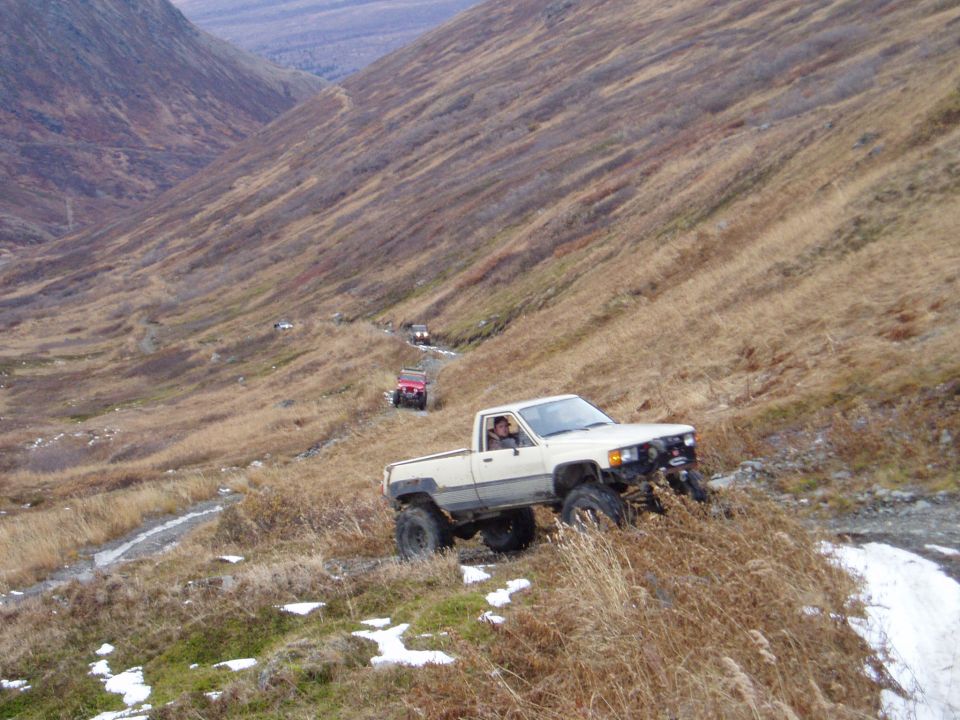 Several off-road vehicles, including a white pickup truck, navigate a dry, grassy mountain trail with scattered snow patches