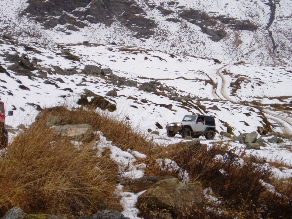 White Jeep Wrangler driving on a snowy mountain trail, another red vehicle partially visible on the left