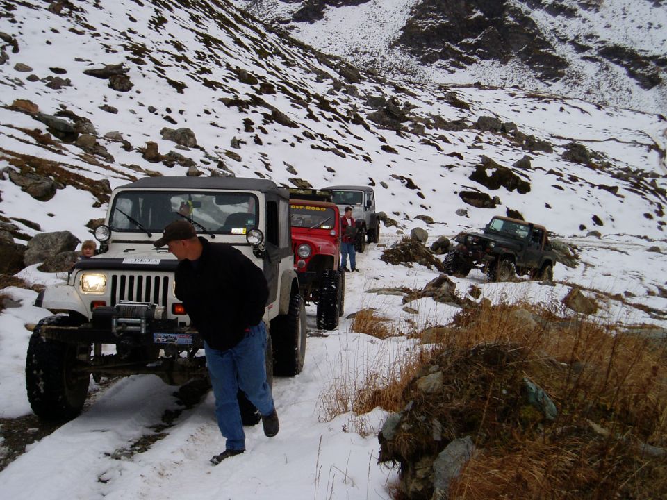 Several Jeeps driving in a line on a snowy mountain path. One person walks beside the lead Jeep