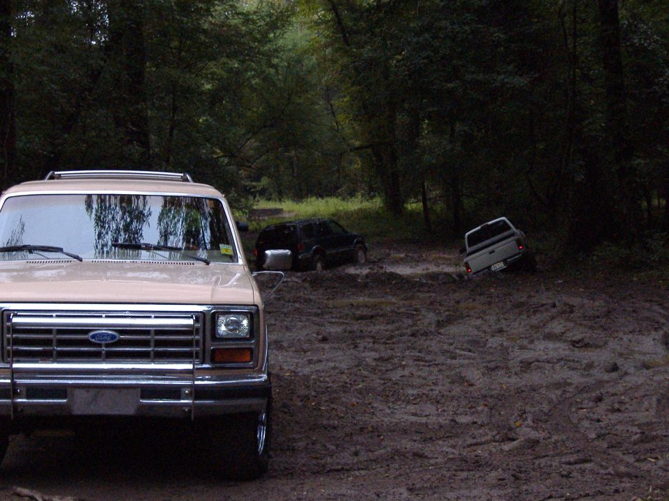 Tan Ford Bronco front end in foreground, two other vehicles stuck in mud behind it on a wooded trail