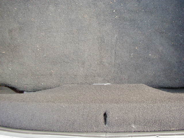 Car trunk interior with gray carpet, showing a small tear and dust. A white line is visible on the upper carpet
