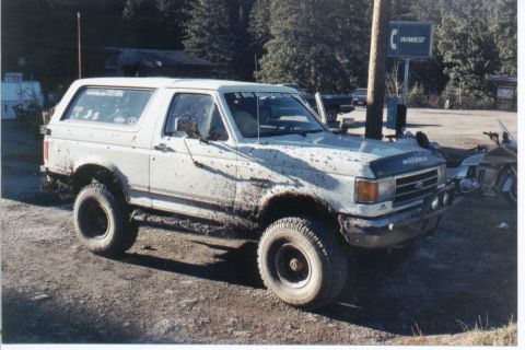 1990 Ford Bronco - Mud-covered 1990 Ford Bronco parked near a pole and trees.