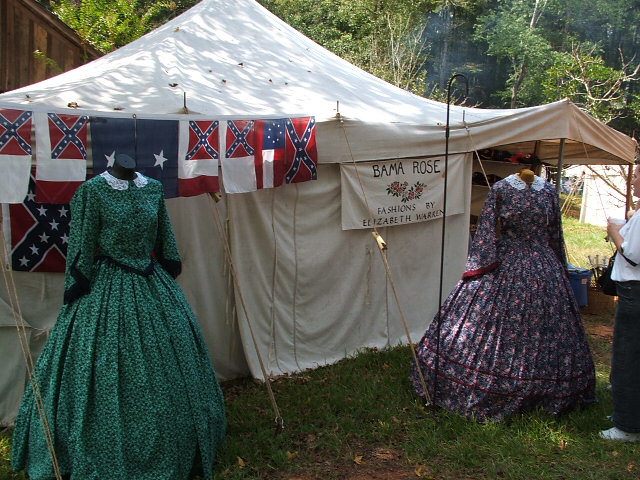 Tent display with two historical dresses and Confederate flags. Sign reads 'BAMA ROSE FASHIONS BY ELIZABETH WARREN'