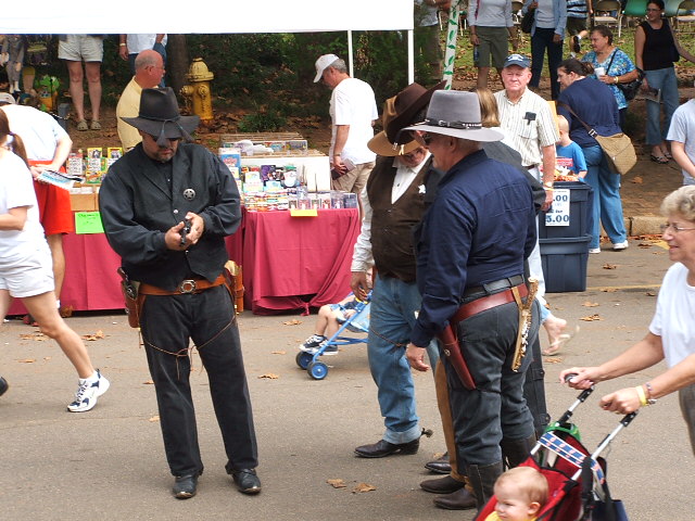 People in cowboy attire stand near tables with merchandise, one man holds a prop gun