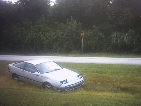 1991 Ford Probe - A 1991 Ford Probe sits in a grassy area beside a road with trees in the background.