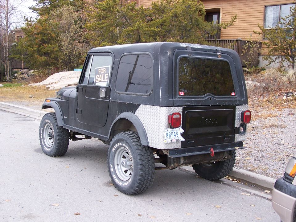 Black Jeep CJ7 hardtop with 'FOR SALE' sign, diamond-plate panels, big mud tires, parked outside.