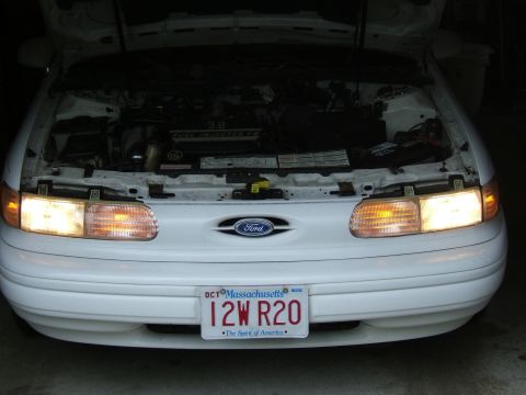 1998 Ford Taurus - The front of a white 1998 Ford Taurus is shown with its hood open, revealing the engine compartment.