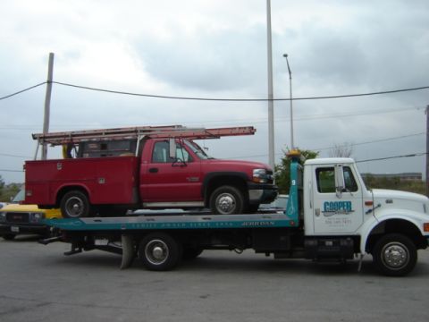 1992 Ford Bronco - A red pickup truck is being towed by a white tow truck. A yellow vehicle is partially visible.