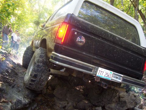 1986 Ford Bronco - Low-angle rear view of a black and white two-tone Ford Bronco SUV climbing over rocks on a wooded off-road trail, showing large mudded knob…